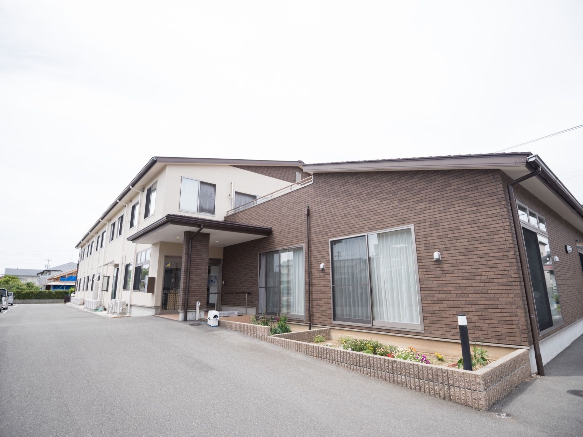 2階建ての白い外壁の建物と屋上のある1階建ての茶色の建物が繋がって建てられている。施設前の路面は舗装されている。