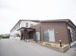 2階建ての白い外壁の建物と屋上のある1階建ての茶色の建物が繋がって建てられている。施設前の路面は舗装されている。