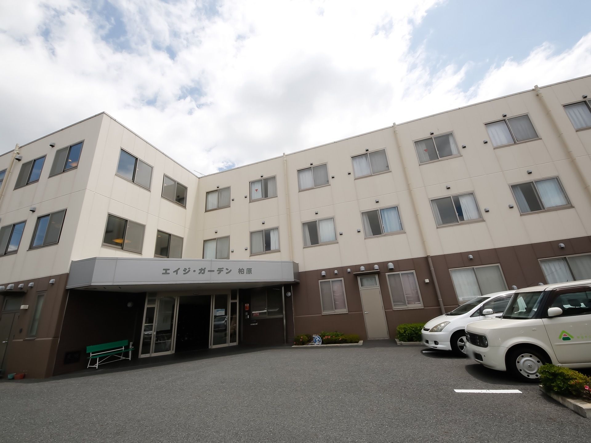 白とダークブラウンの外観の建物である。Ｌ字型の角に玄関を設け、屋根の側面に施設名を表示している。敷地内には駐車場スペースを備えている。