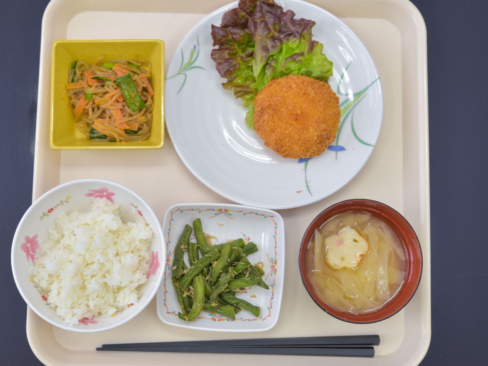 一人分の食事がお盆の上に乗っている。白米と麩入りのお味噌汁に、おかずはコロッケ、インゲンのじゃこ和え、煮物の小鉢もついている。