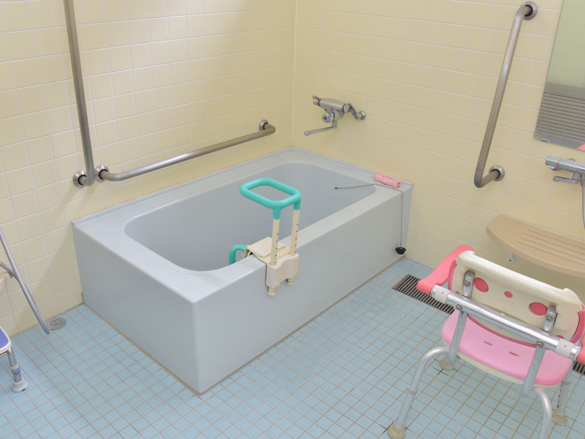 お風呂の浴槽には、介護用の入浴介助機器が設置されている。洗い場と浴槽両方に手すりが付いていて、シャワーチェアが置かれている。