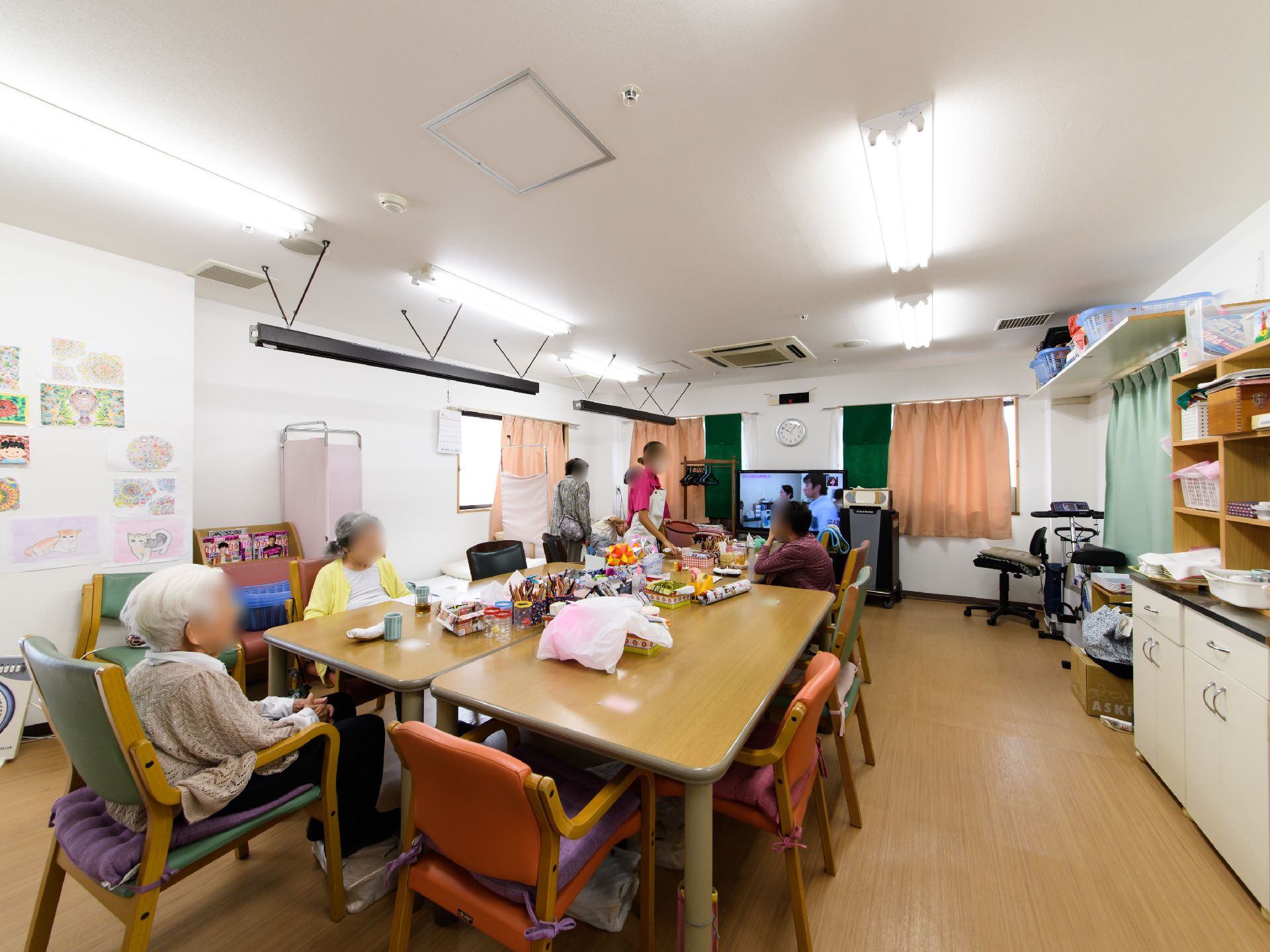 広い室内には、テレビやテーブル、椅子、ベッドが置かれている。壁には利用者様が描いたと思われる絵が飾られている。