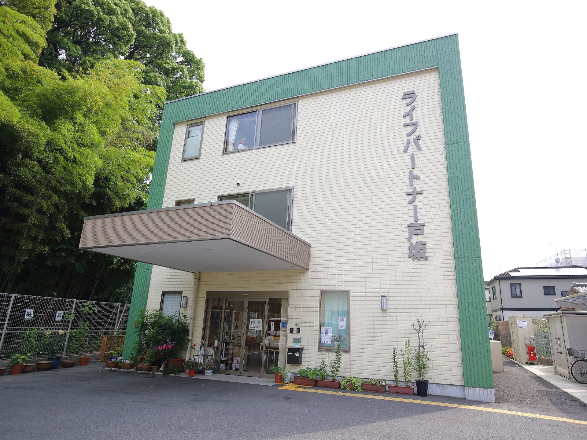 三階建て、明るいベージュ色と緑色に縁どられた建物。。出入り口には雨除けがある。多数の植木鉢が施設を彩る。