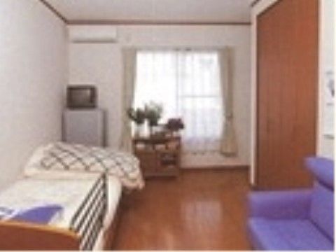 フローリング敷きの小さめの部屋で、奥に大きな窓が付いている。ベッドやテレビ、冷蔵庫など家具が一通り揃っている。