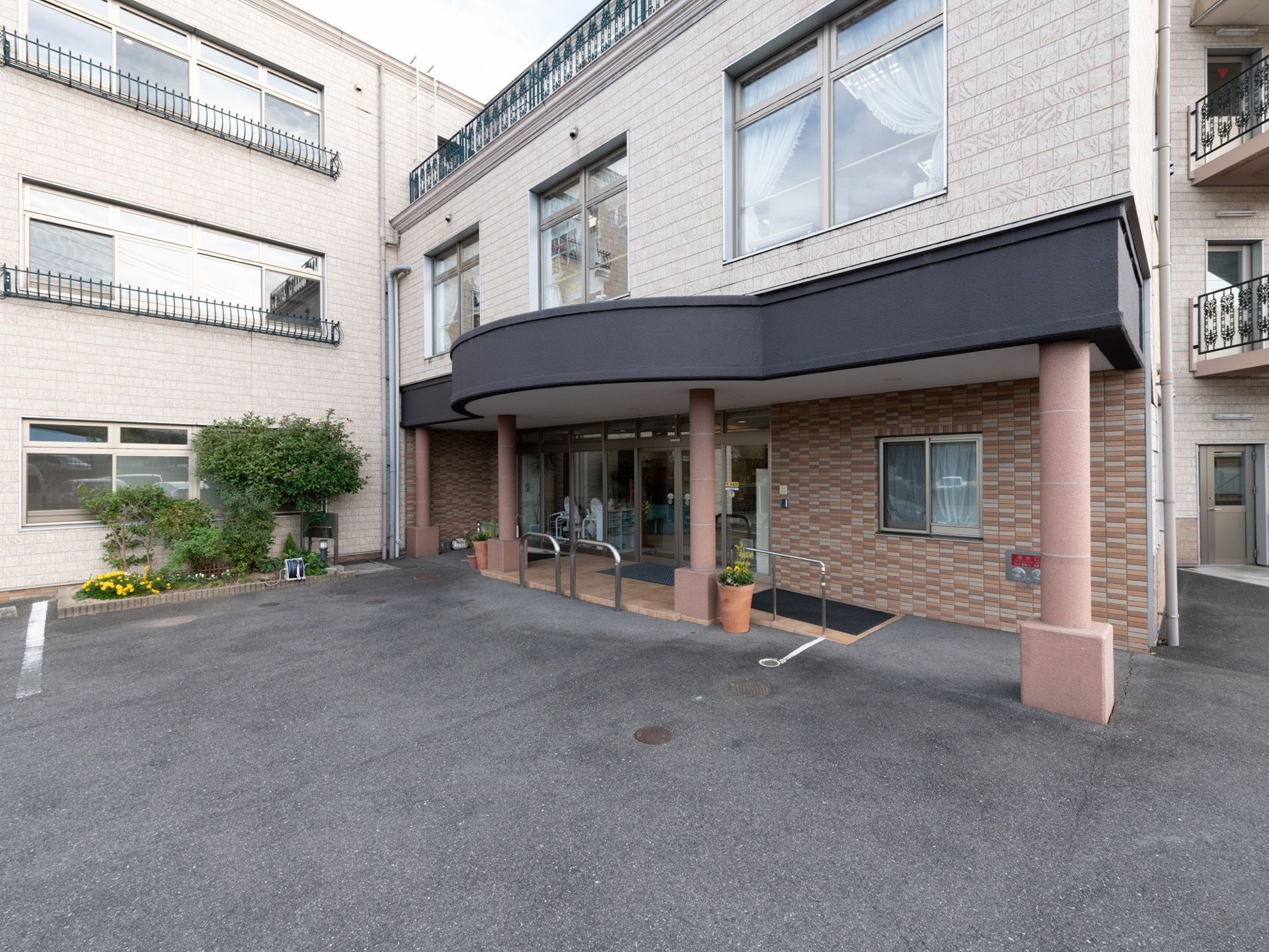 ベージュの外壁の３階建ての建物である。出入り口には丸い柱が４本あり、手すりとスロープが設置されている。