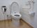 施設のトイレはウォシュレットの水洗トイレ。壁には手すりとスイッチ、インターホンがある。便器の横には可動式の手すりも設置されている。