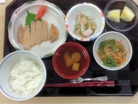 黒いトレーの上に、主食の白ご飯、主菜、副菜、フルーツなど６品が並んでいる。赤い塗り箸と小さなフォークが添えられている。