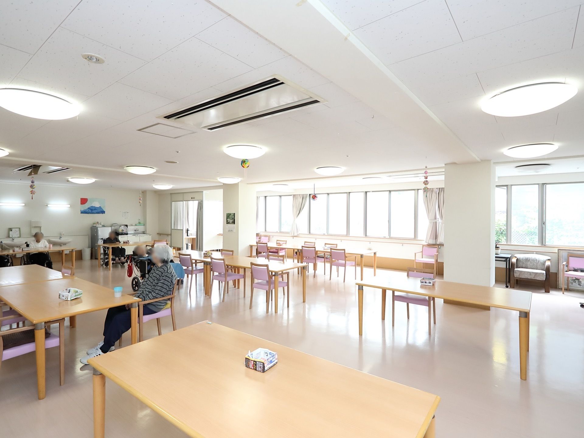 テーブルが10台以上並べられたリビングがある。壁には手すりがつけられていて、洗面台や冷蔵庫が設置されている。