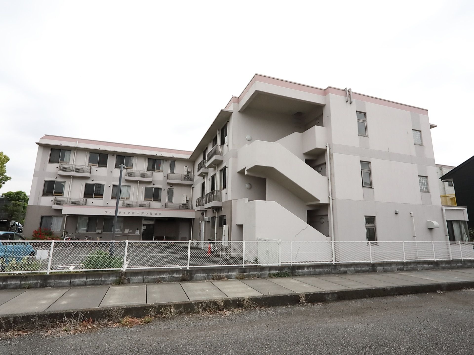 3階建てでL字型をした白い建物がある。敷地は背の低いフェンスで囲まれている。建物の前は広い駐車スペースになっている。