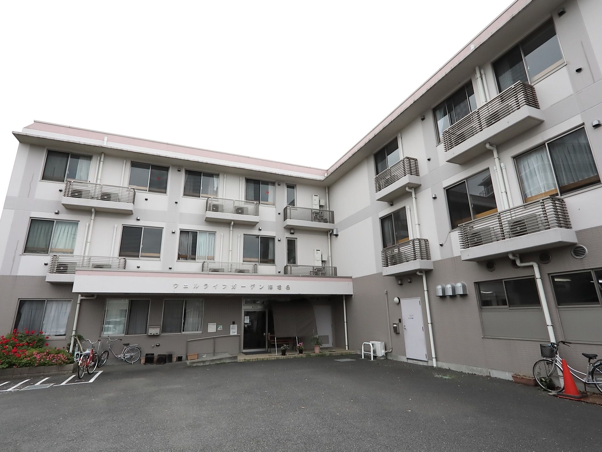 L字型で3階建てのマンションタイプの建物。1階部分の壁はグレーで、2階以上は白くなっている。建物の脇には駐輪スペースがある。