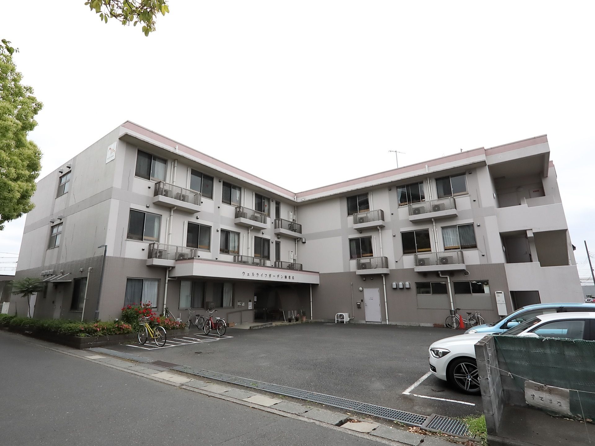 白とグレーの2色が印象的な3階建てのマンションが建っている。敷地内は舗装されていて、駐輪場や駐車場が整備されている。