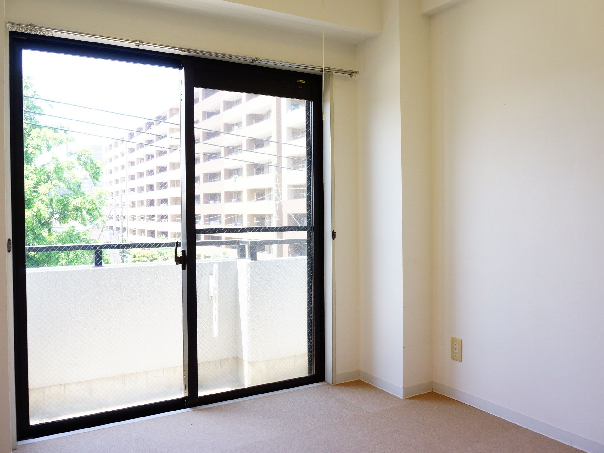 家具などが何も置いてない洋室の部屋で、カーテンレールが取り付けてある。壁は白い色である。窓は吐き出し窓で、ベランダがついている。