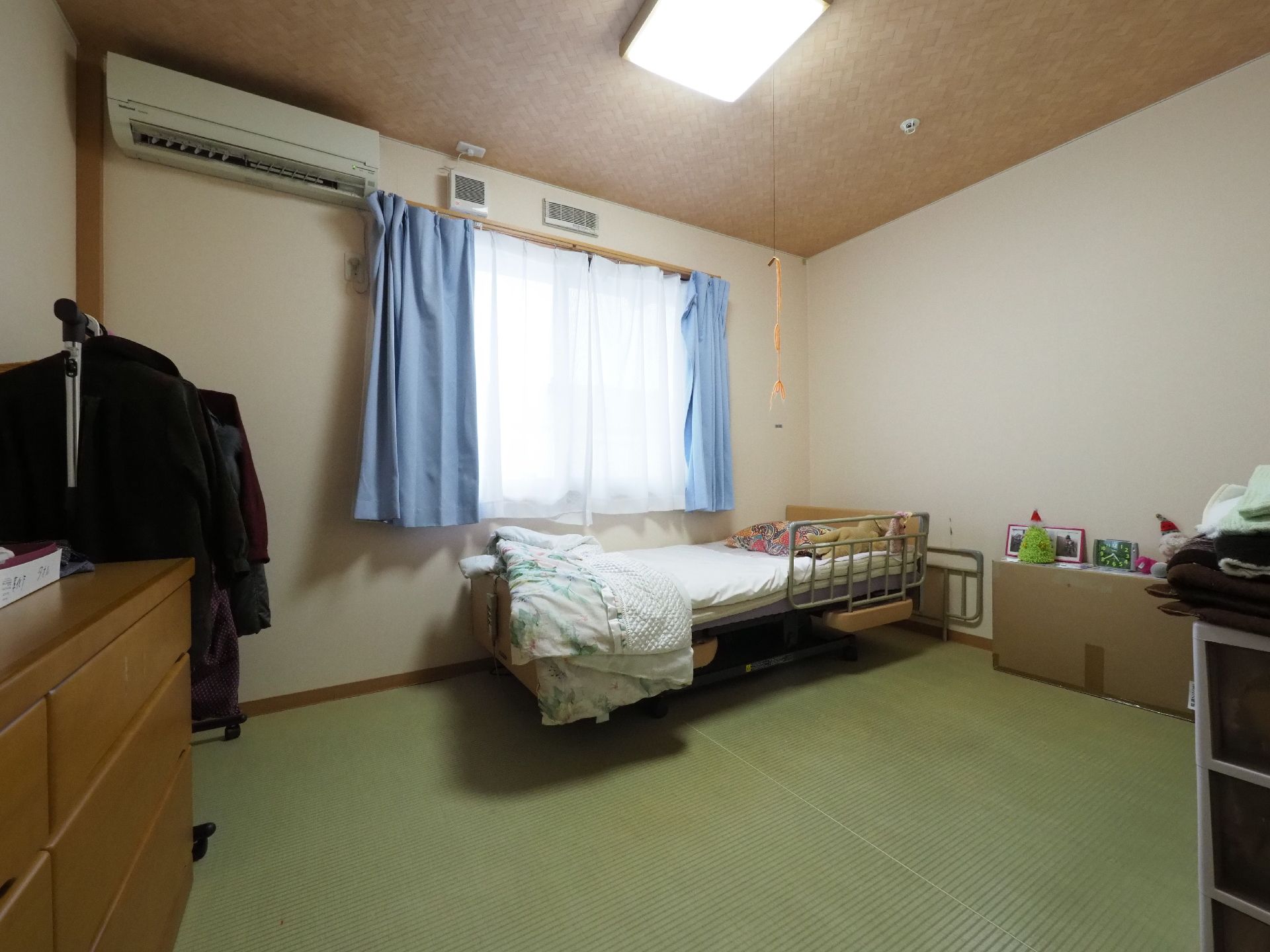 畳敷きで介護用ベッド、洋服ダンス、パイプハンガーが置かれ、腰高窓がある居室