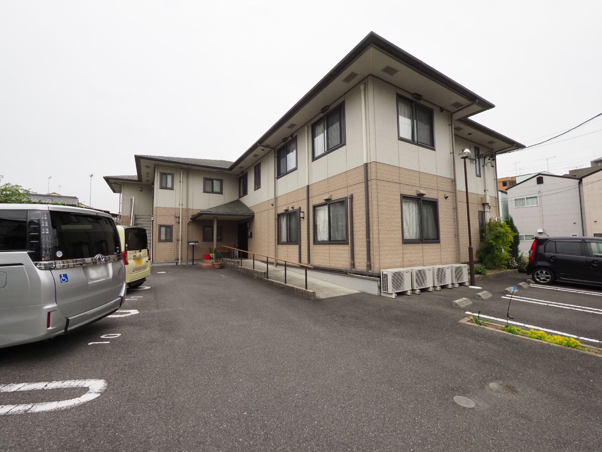 L字型の駐車場と、エントランスへのスロープが設けられた２階建ての施設の外観