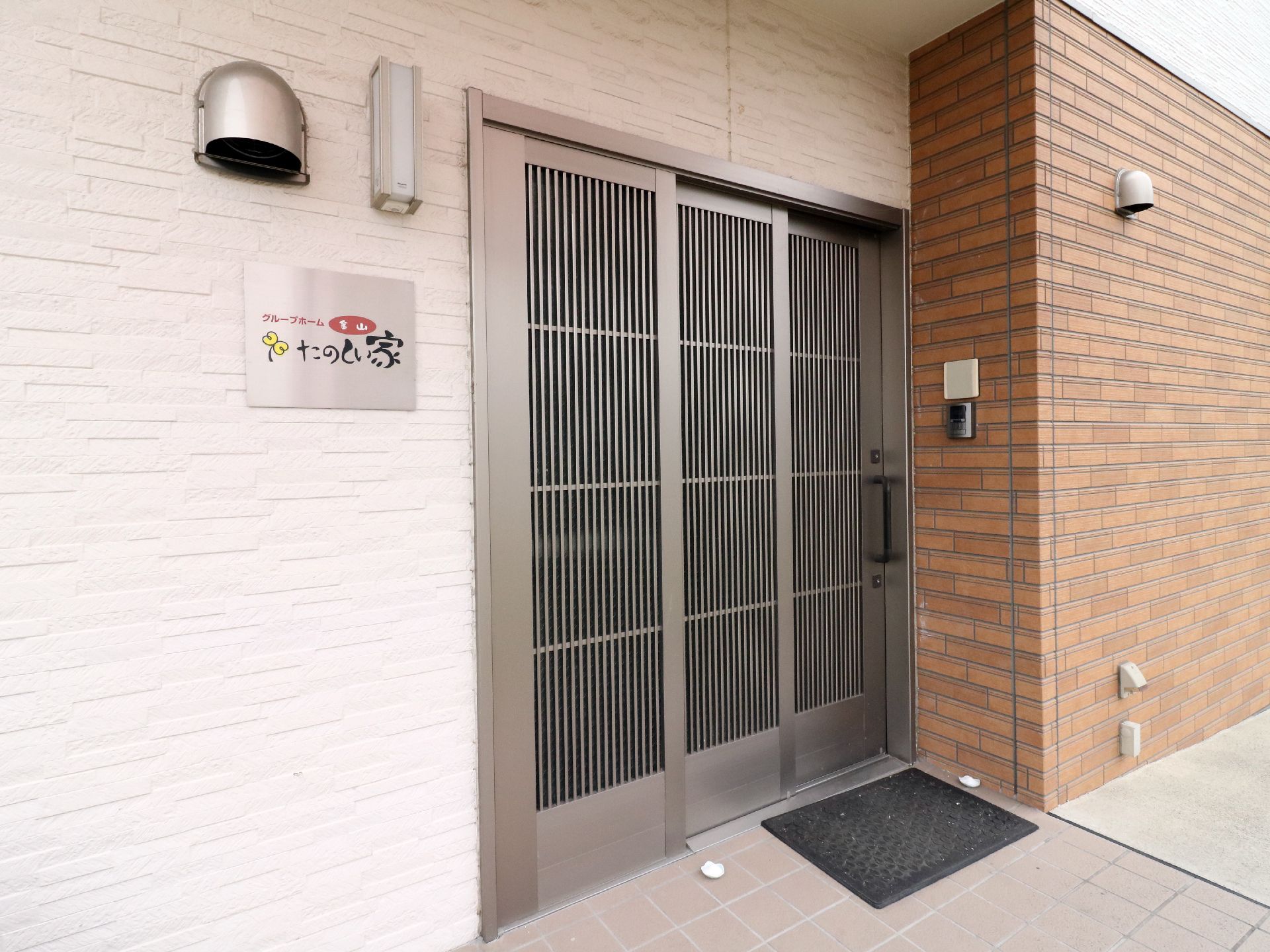 施設の玄関は金属製の引き戸になっていて、ドアの周りにはインターフォンや、同じく金属製の表札が設置されている。
