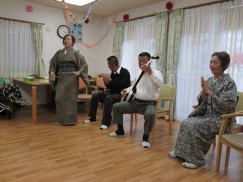 着物を着て歌う女性と、椅子に座りながら楽器を演奏する男性