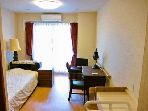 掃き出し窓に赤いカーテンとレースカーテンが掛けられている。ベッド脇に小さなチェストが置いてあり、壁際にデスクと椅子が配置されている。