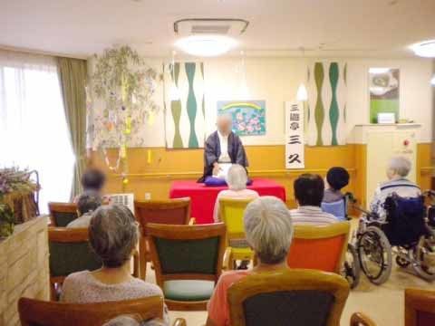 正面の赤い台座に座っている落語家の話を聞いている入居者たち