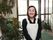 エントランスで撮影された女性スタッフの写真。黒い襟付きのポロシャツと施設名が書かれた白いエプロンを着用している。