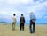 海岸を散策中の３人の男性の後ろ姿である。白い雲が流れる空の下で、２人は海に向かって立ち、１人は歩きながらそれぞれ景色を満喫している。