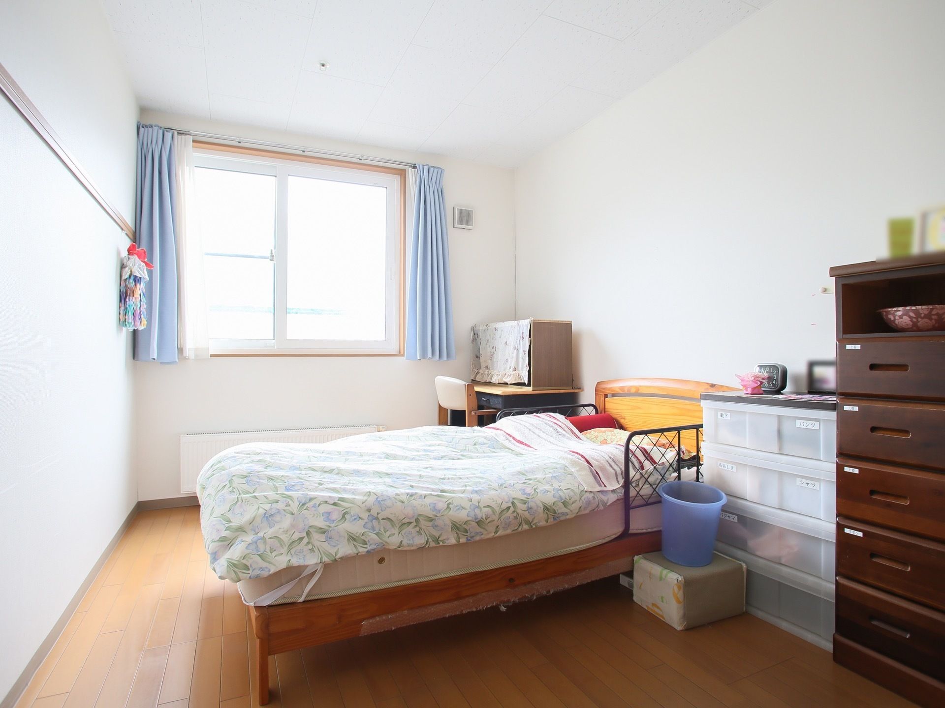 大きな窓から外光が照らす明るい居室である。ベッドや机、収納タンスが置かれている。壁には館内放送用のスピーカーが設置されている。