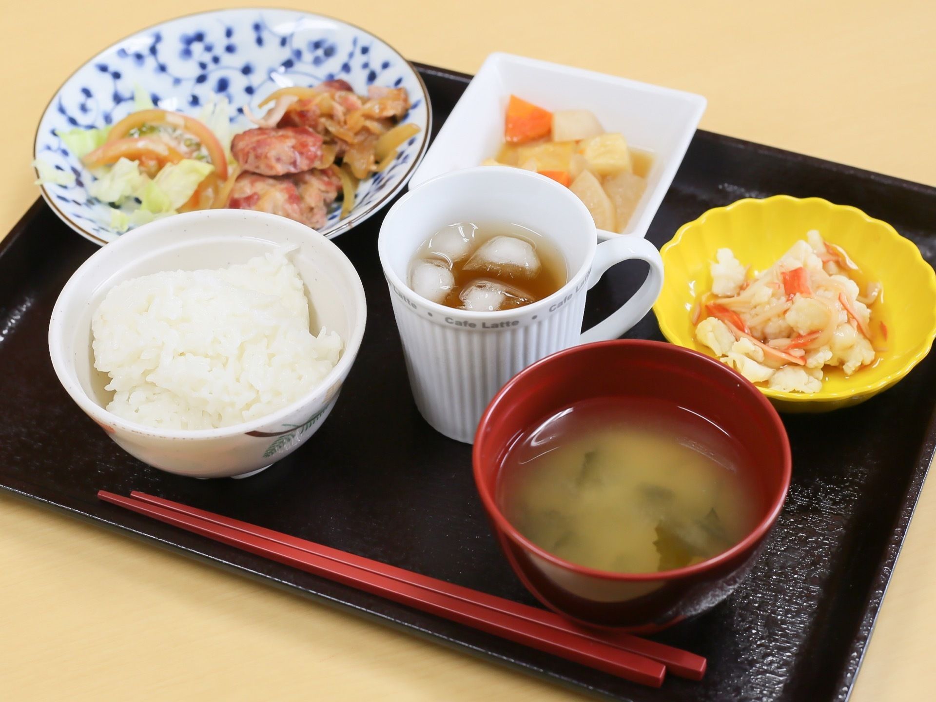 黒色のお盆上に、ご飯とお味噌汁、ほか数品目のおかずや飲み物が並べられている。おかずは野菜を中心とした具材が用いられている。
