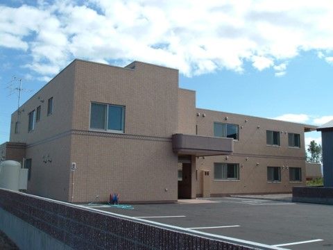 ベージュ色の外壁をもつ、コンクリート素材のシンプルな建物。２階建てで玄関にはひさしが作られており、建物の前には駐車スペースがある。