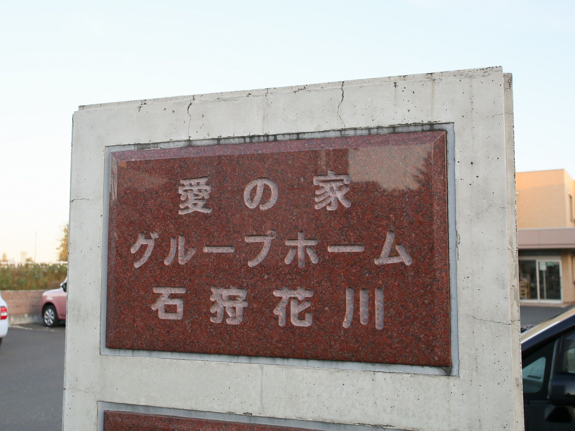 アスファルトで舗装された駐車場前に立てられた看板。施設名が書かれた重厚感のある茶色いパネルの表札となっている。