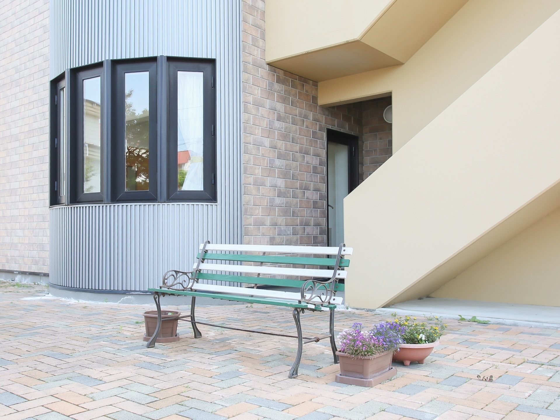 奥にレンガ張りの施設があり、外階段が見えている。手前は石畳になっており、白と緑のベンチが置かれている。