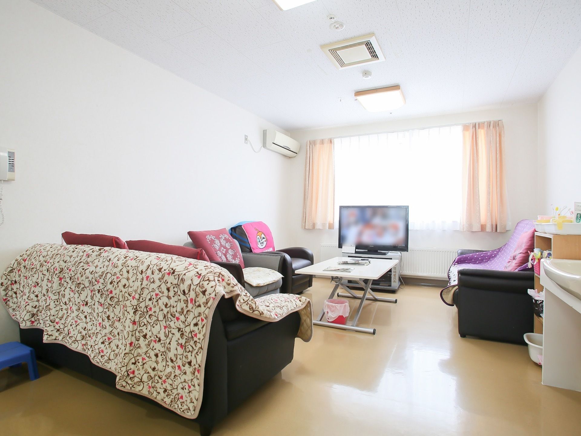 白い壁の共有スペースにたくさんのソファが置かれ、座布団などが敷かれている。窓の前に大型液晶テレビが用意されている。