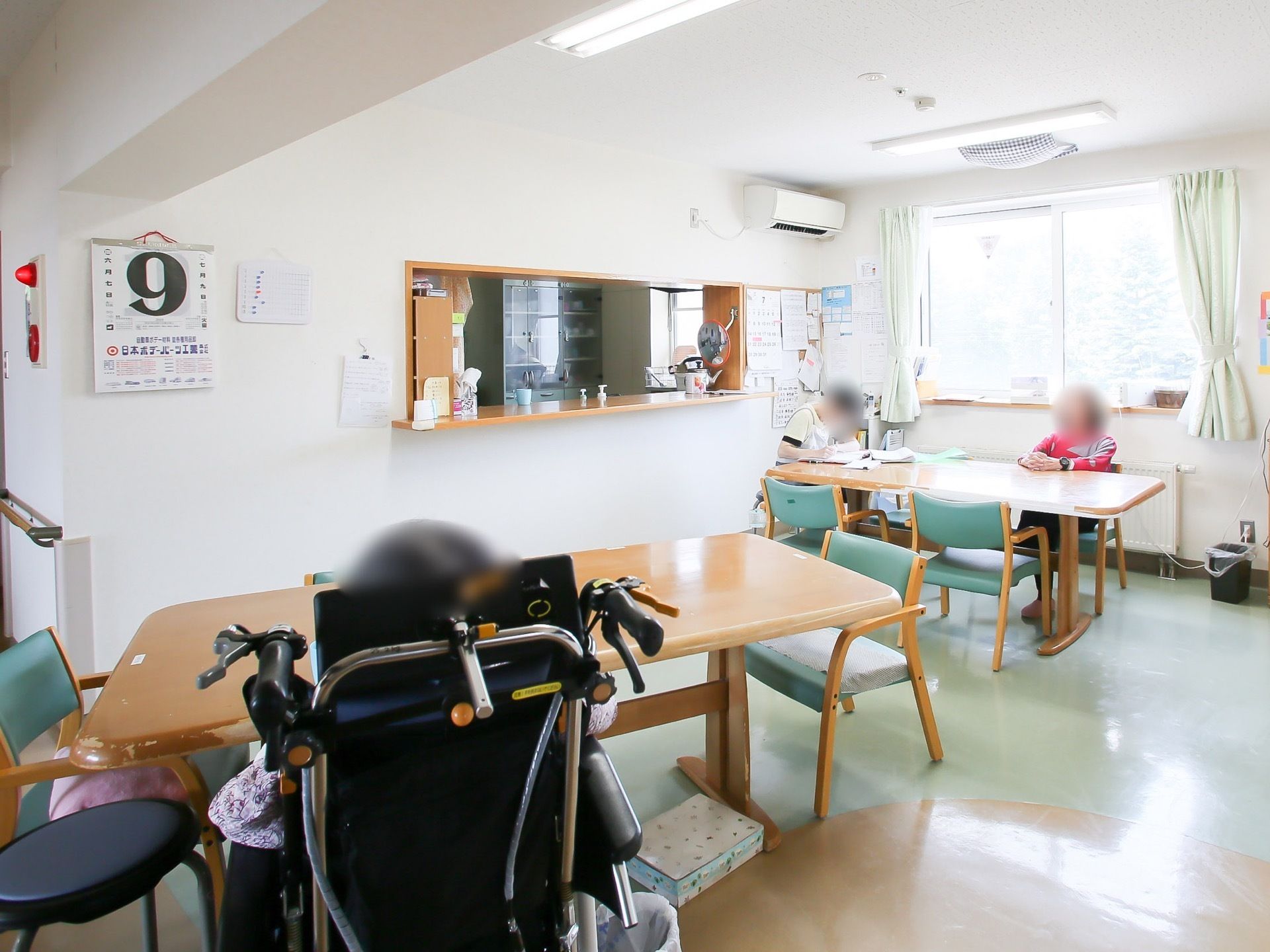 正面にキッチンへ続く窓があり、その横の壁にコルクボードが設置されている。手前にはたくさんのテーブルセットが並んでいる。