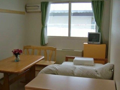 引き違い窓の外には白い格子状の柵が付いている。テレビを一つ置き、木製のベッドにはテーブルがつけてある。