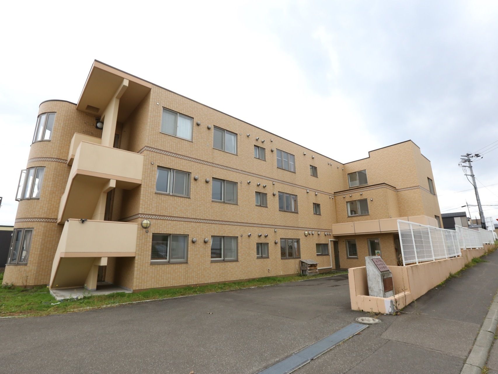 住宅街の中にあるブラウンを基調とした落ち着いた色合いの建物である。歩道との間には、白いフェンスが立てられている。