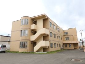 オレンジ色のタイル調の壁が特徴の建物である。非常階段横の出窓がオシャレである。建物の周りは駐車スペースとなっている。