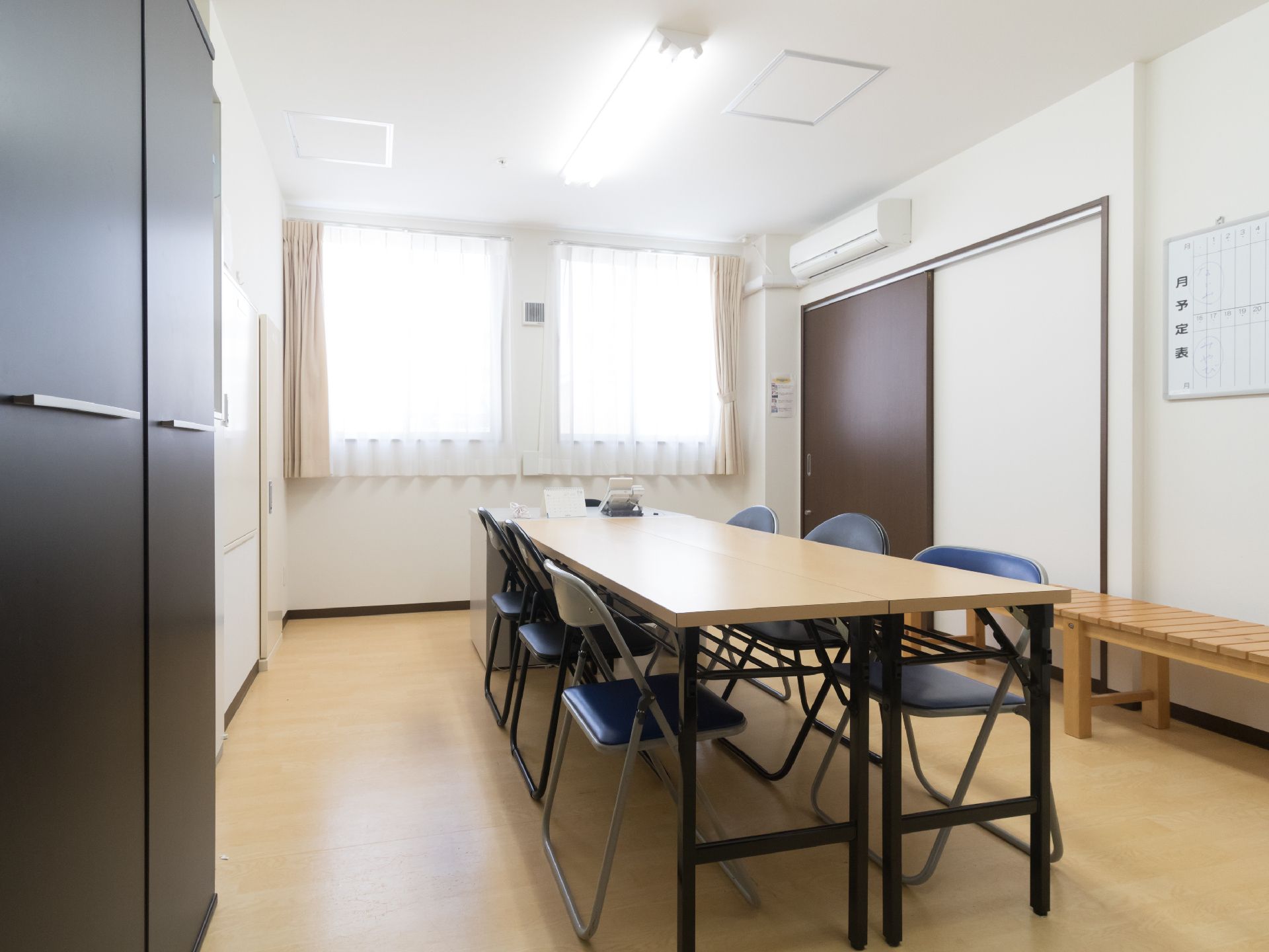 部屋には6人掛けの長机と椅子が置かれている。壁にはエアコンや月予定を書き込むホワイトボードが取り付けてある。