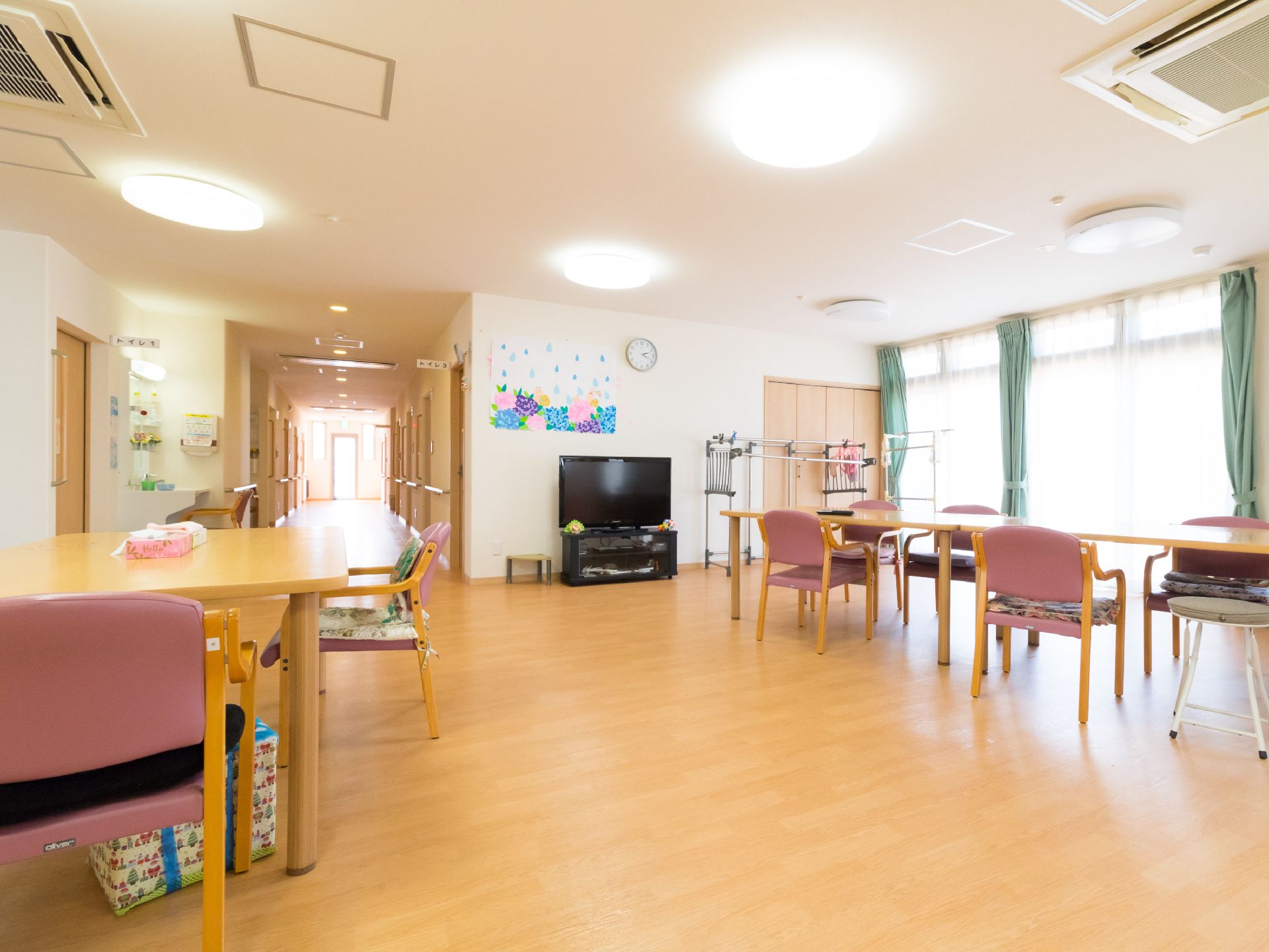 テレビが置かれた部屋がある。茶色いテーブル、肘置き付きのピンクの椅子も複数並べられている。壁には丸い時計がある。