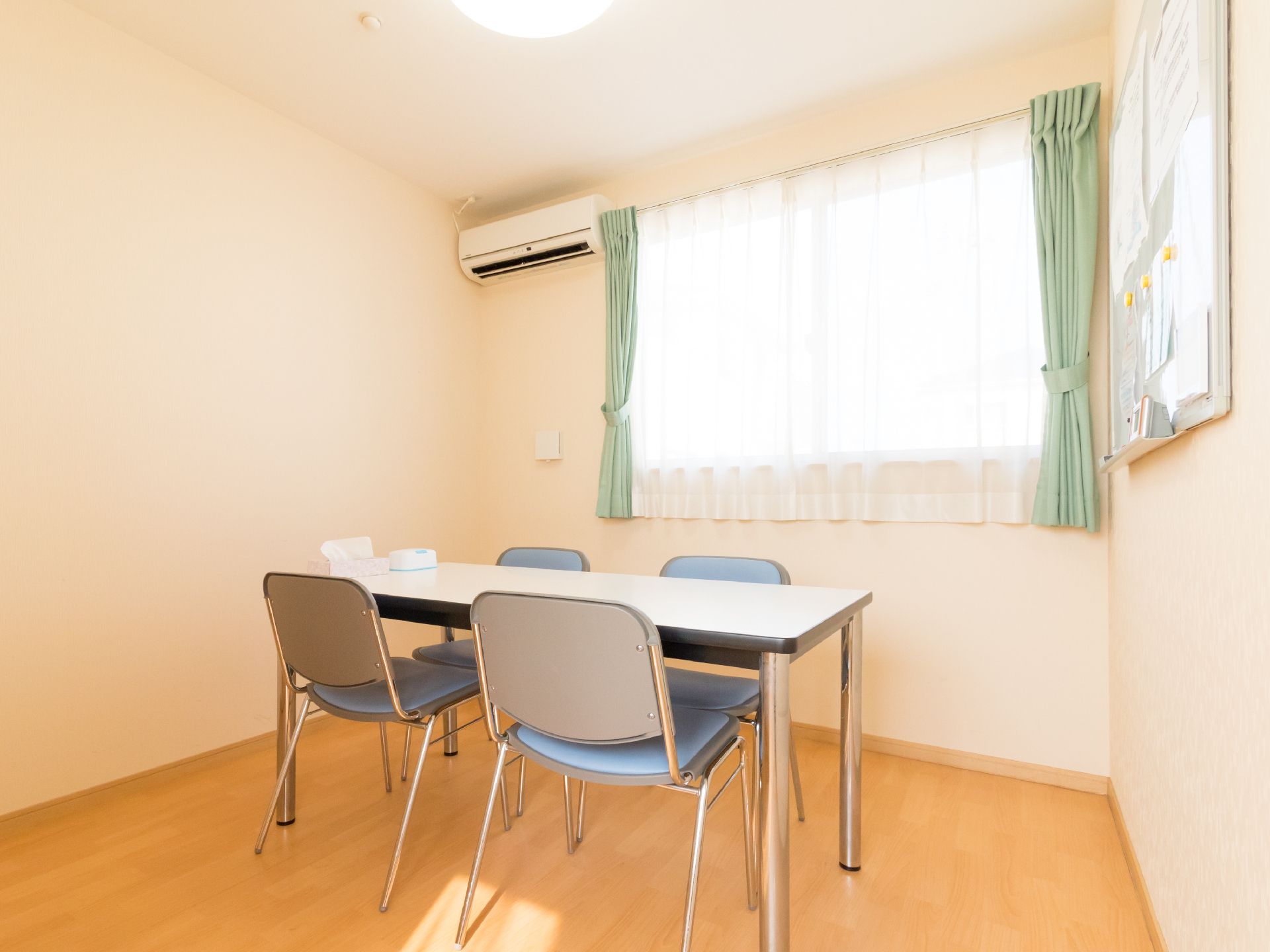 レースのカーテンと黄緑色のカーテンが掛かった部屋には、白いテーブルと青いパイプ椅子が4脚並べられている。