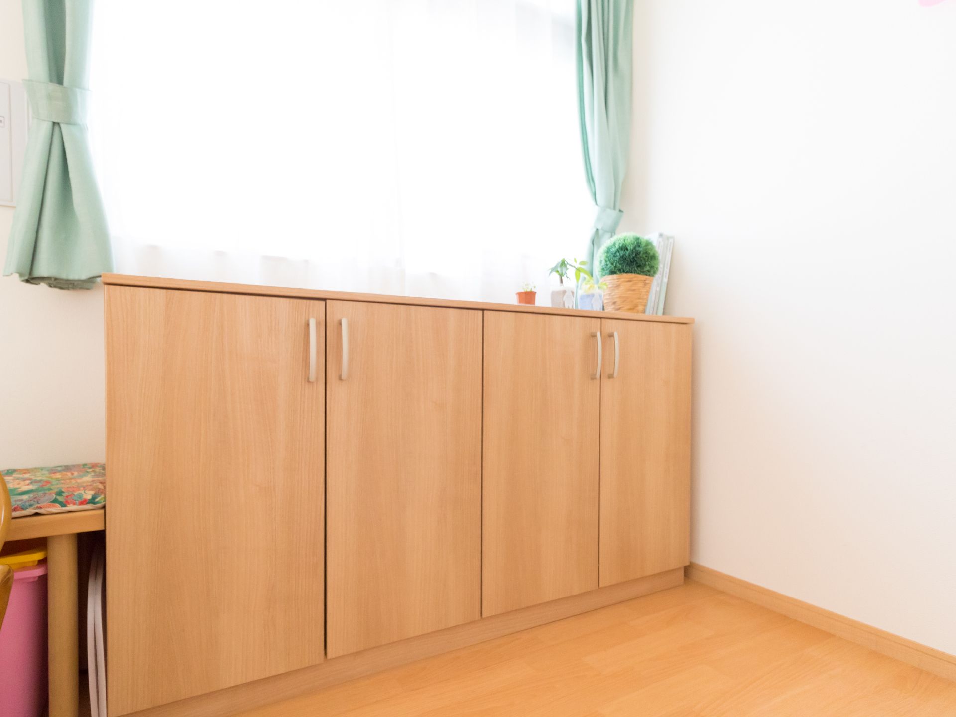 レースと緑色のカーテンが掛かった窓が設置された部屋がある。窓際には両開きの扉が2つある棚が置かれている。