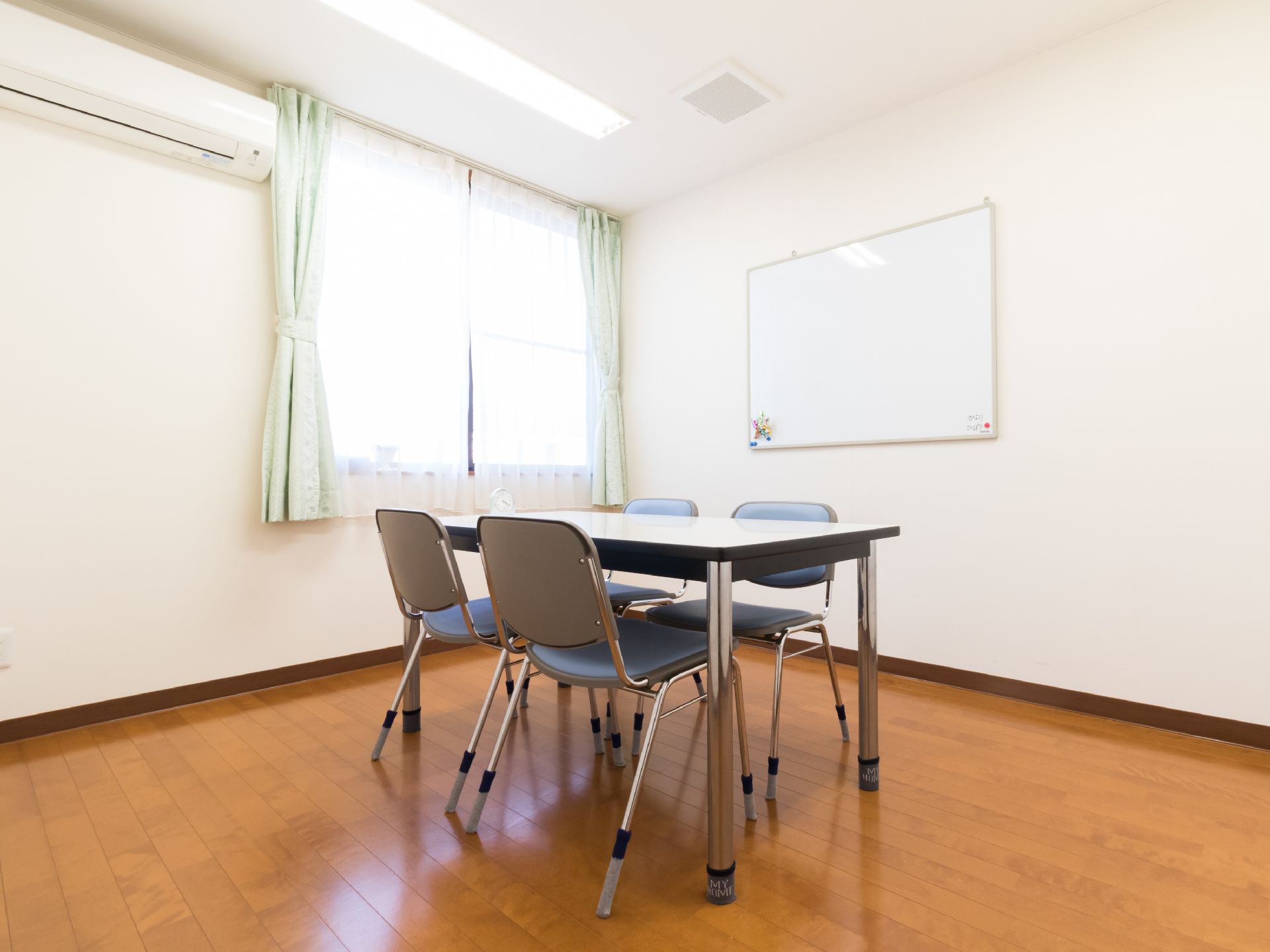4人掛けテーブルと椅子が余裕を持った広さの部屋に配置されている。壁にはホワイトボードとエアコンが設置されている。
