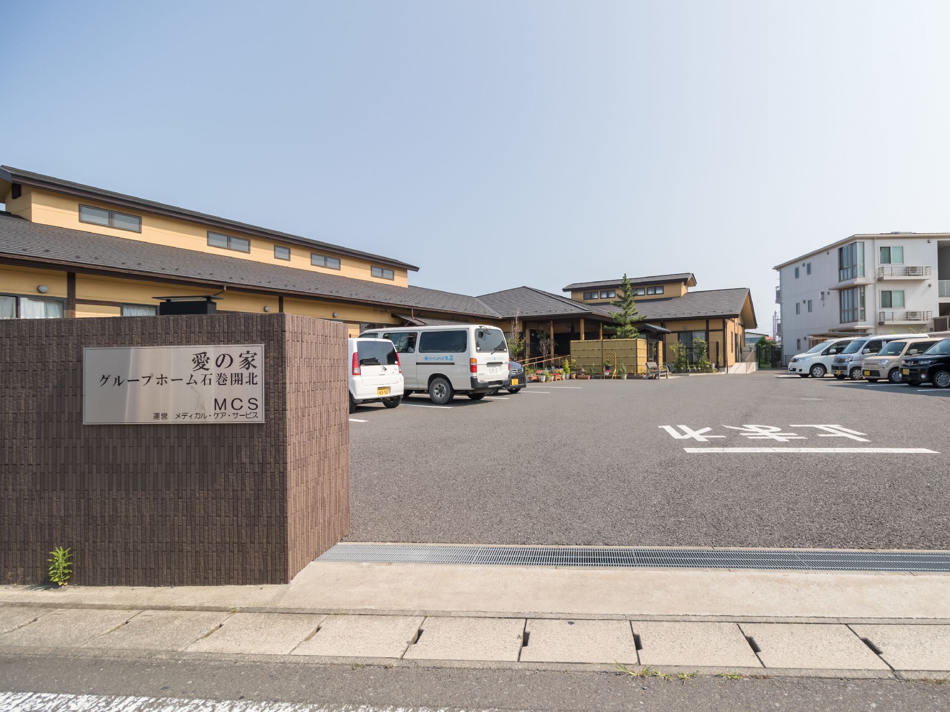 敷地内入口の塀には施設の名前が書かれたプレートが付けられている。建物の前には広々として駐車場が設けられている。