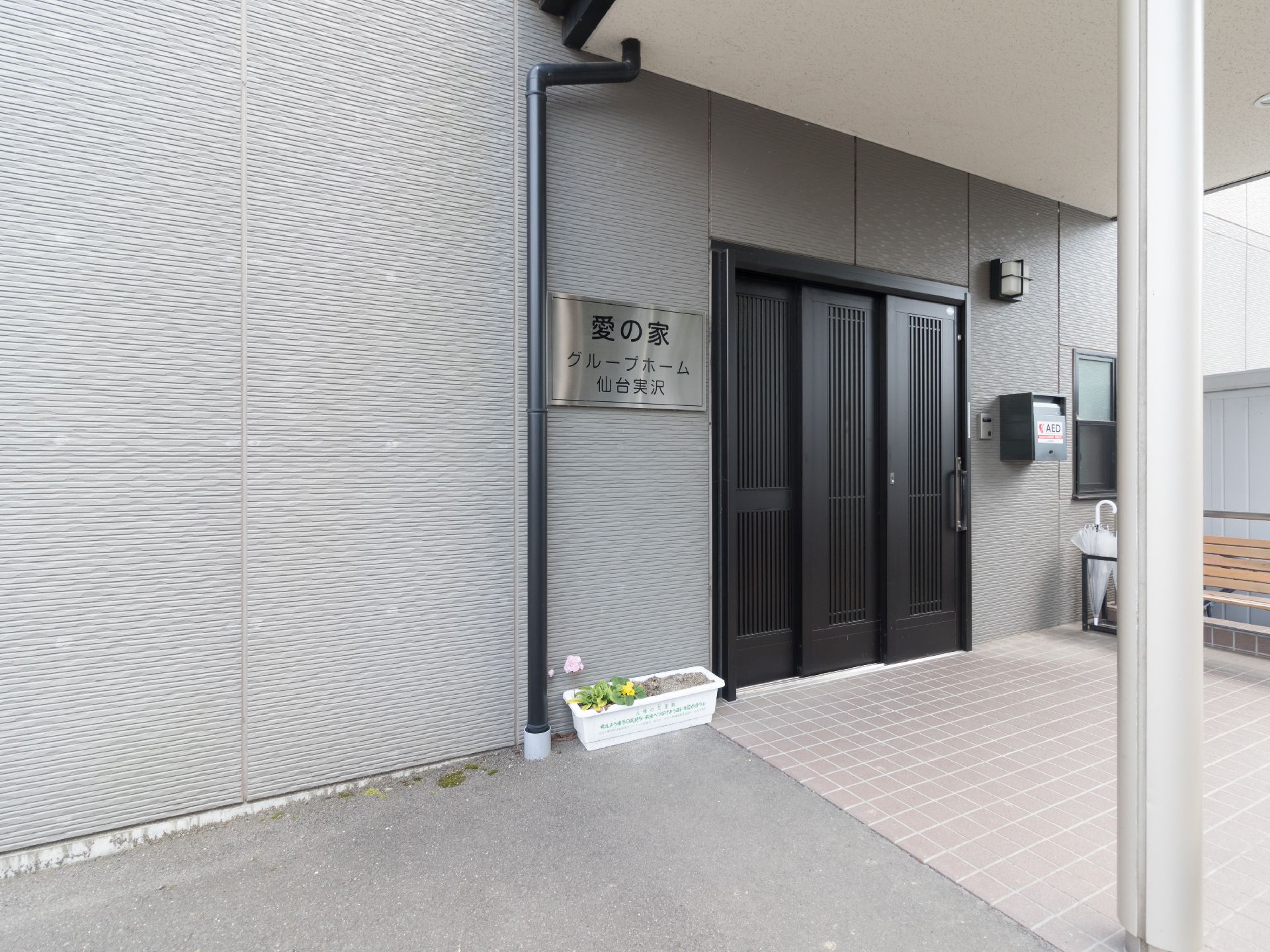 広いスペースが設けられたエントランスにはベンチや傘立てが置かれている。ドア横の壁には施設名の看板と郵便受けが取り付けてある。