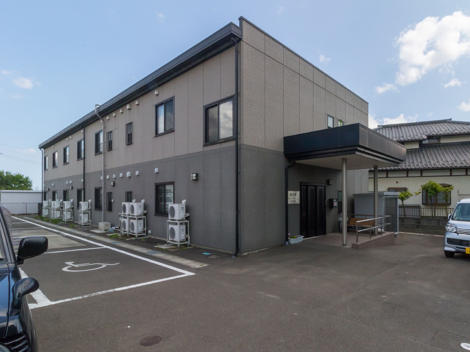 落ち着いたシックな色合いの2階建の施設である。車椅子仕様車も停車できるスペースを設けた広い駐車場が施設前にある。