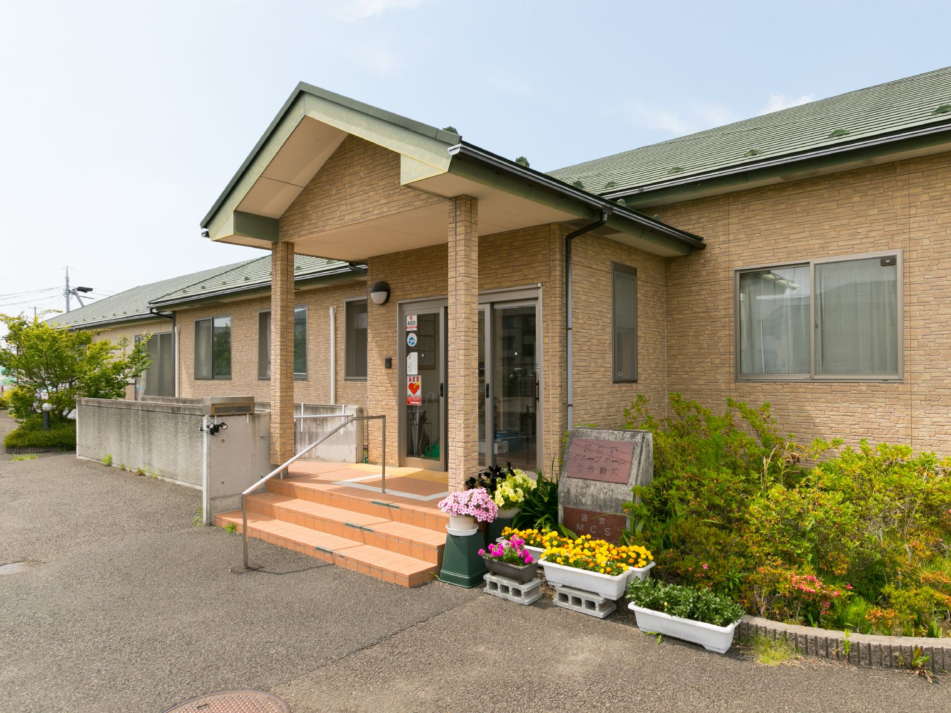 建物の入り口では、黄色やピンクの花がプランターで育てられている。階段を3段上がると、屋根のあるスペースがある。
