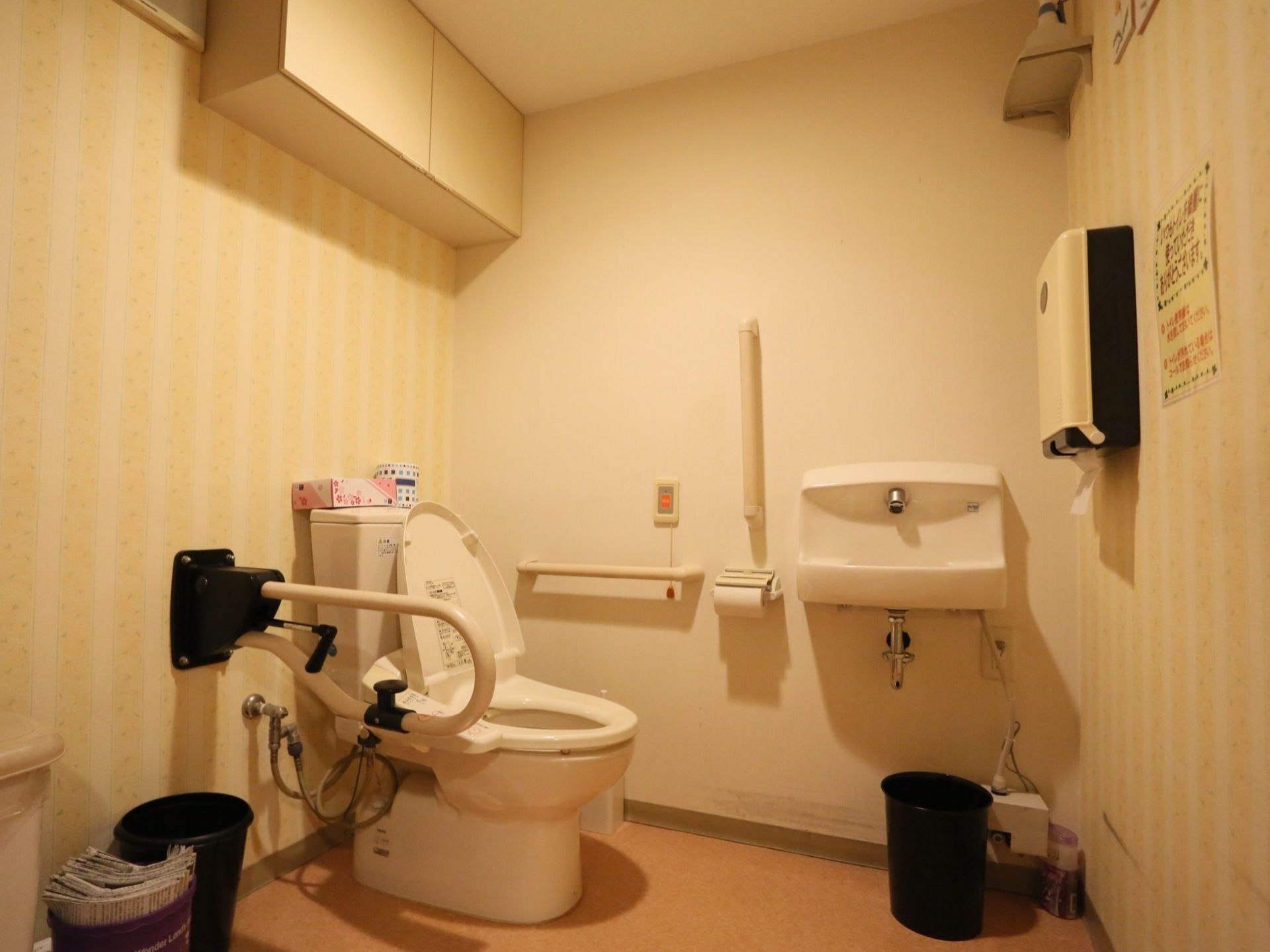 個室の広いトイレがある。壁には手すりがあり、便座の横には可動式のひじ掛けが設置されている。側には洗面台がある。