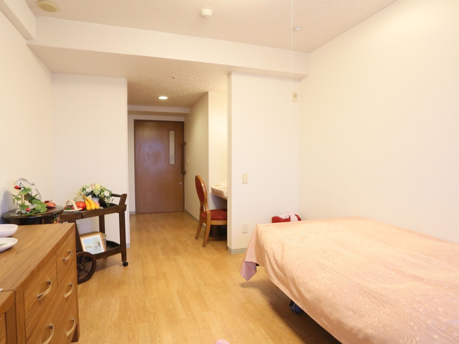 明るいフローリングの居室がある。チェストとワゴン、ベッドが置かれている。入り口付近には洗面台があり、椅子がある。