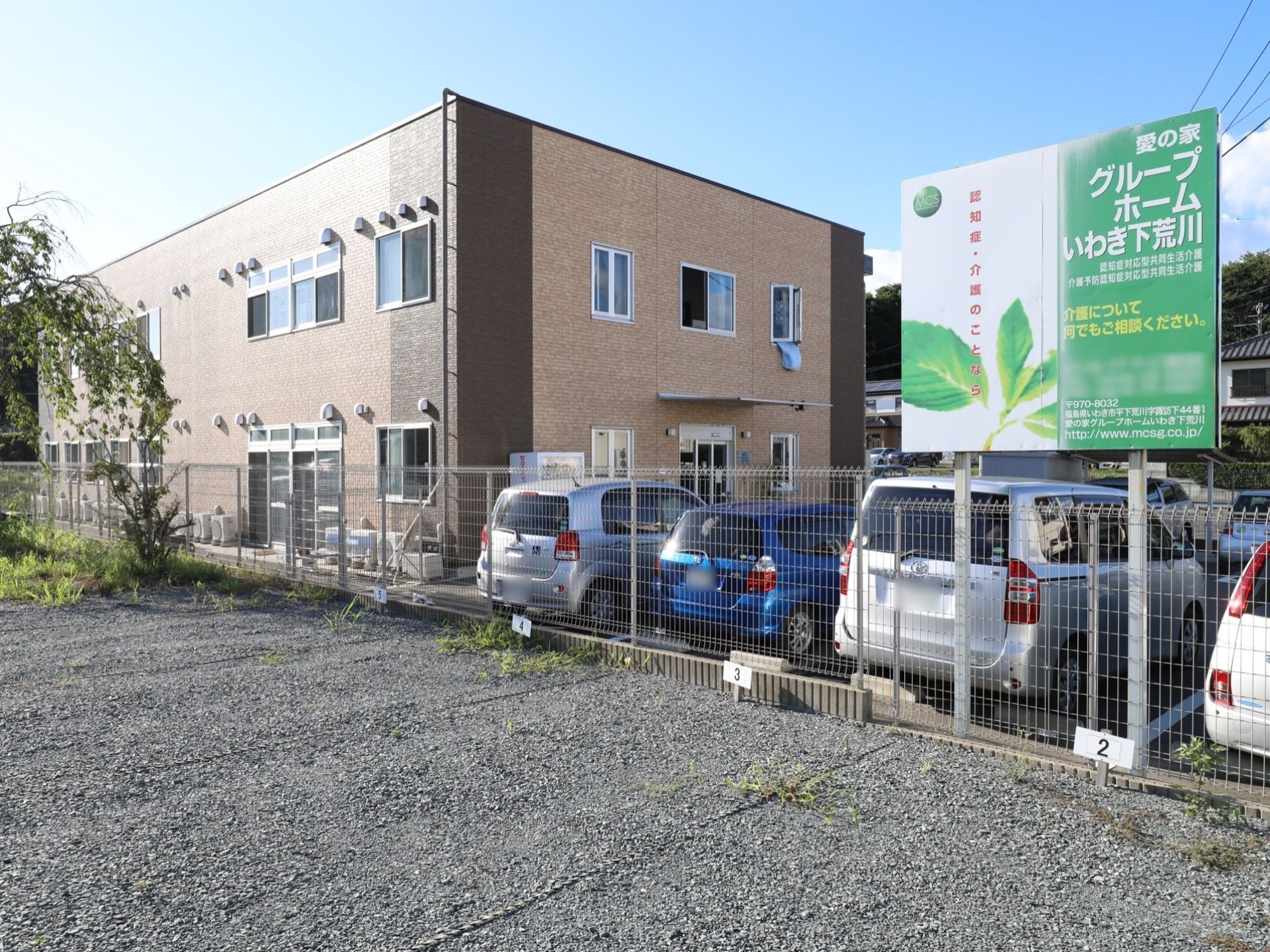 落ち着いた環境の中に建てられたグループホームである。駐車場が完備されており、施設の看板が立てられている。