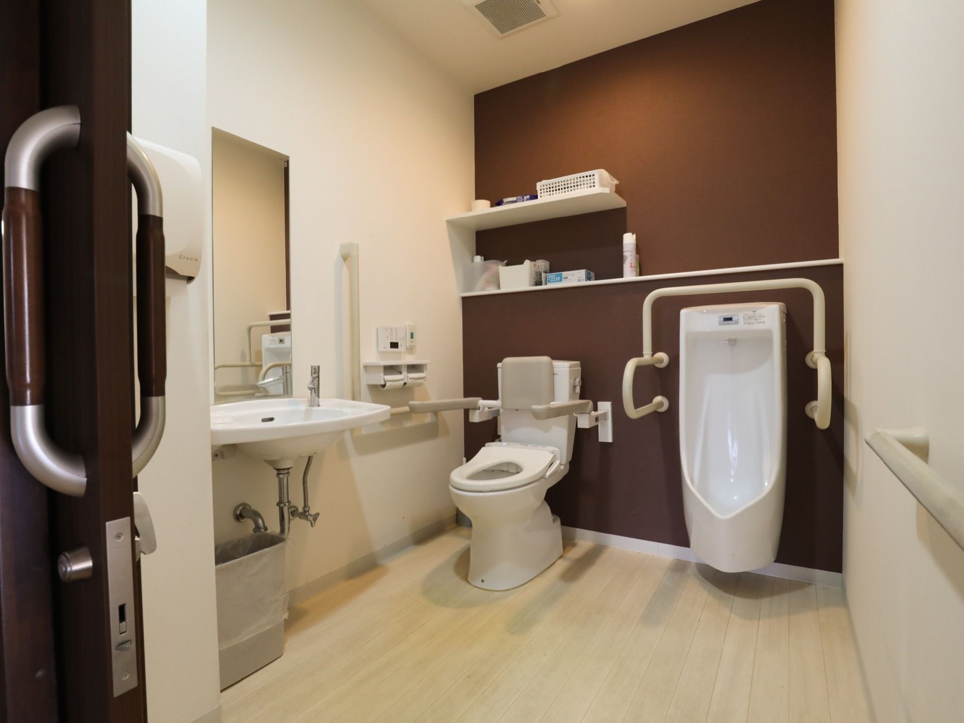 清潔感のある個室トイレで、便座には背もたれやひじ掛けが取りつけられている。壁には棚が設置されている。