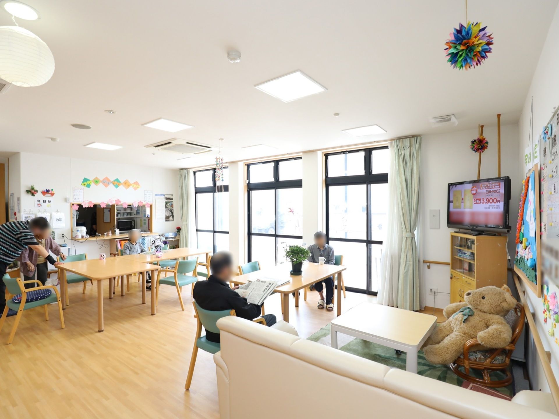 共有スペースでは、テーブルセットの椅子にそれぞれに腰かける男性の入居者様３名の姿と、スタッフに移動の介助を受けている入居者様の姿が見える。