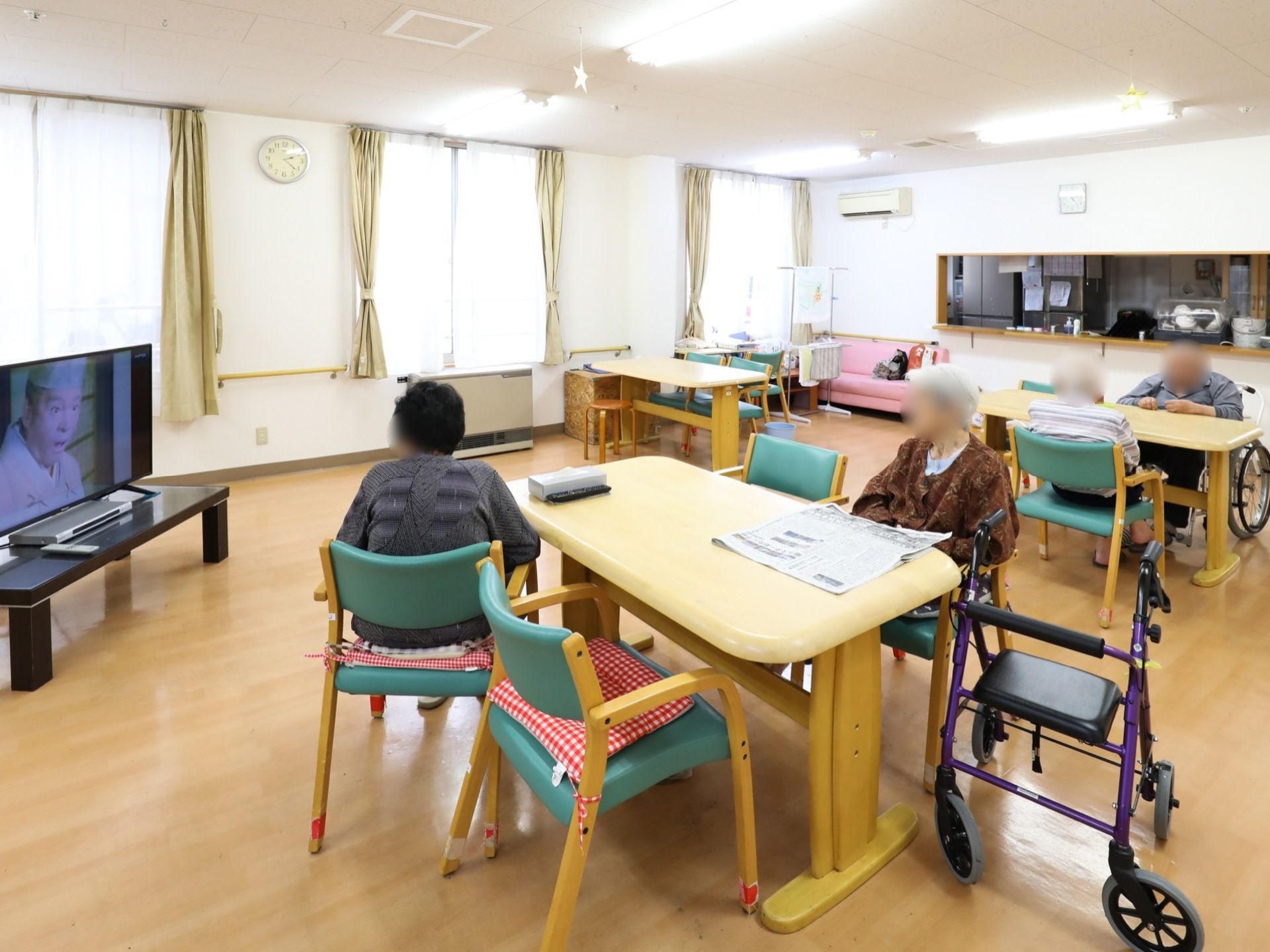 食堂では、車椅子をご利用の方や歩行器を使用している方など数人の入居者様が、テーブルに着席してテレビを見ている。