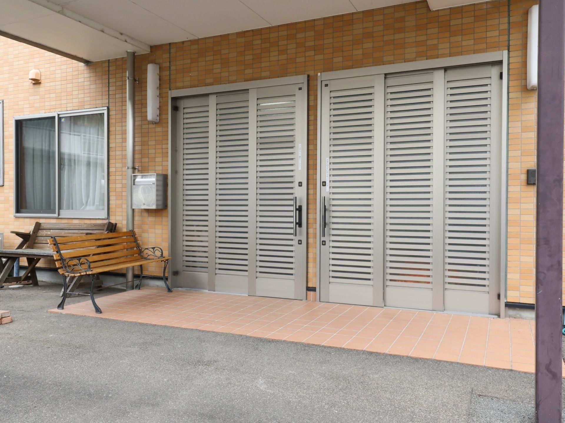 雨除け屋根のついた玄関には、シルバーの３枚扉が左右に並んで設置されている。脇には郵便ポストがあり、ベンチが置いてある。